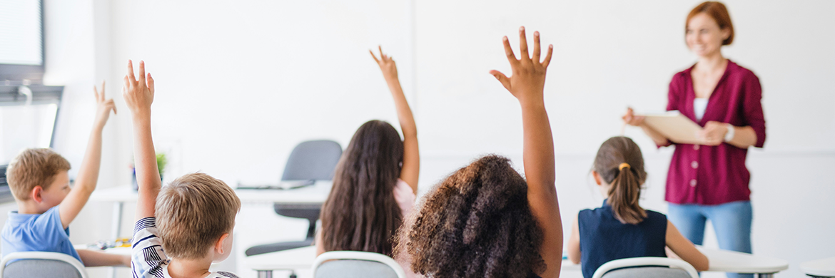 Class of students raising their hands with a female teacher smiling at the front of the class